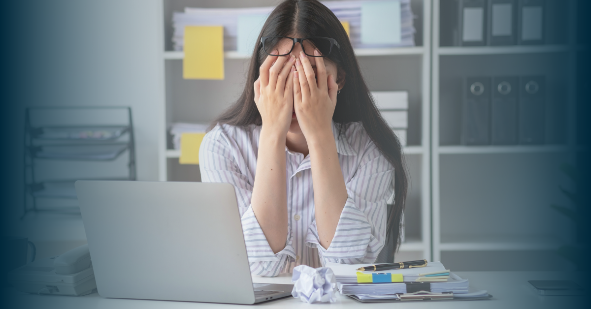 A young woman with long dark hair sits at a desk. There is a silver laptop, clipboard, pens and a screwed up ball on paper on the desk in front of her. She holds her head in her hands, obscuring her face completely, her glasses pushed up to her forehead. She gives an air of stress and frustration.