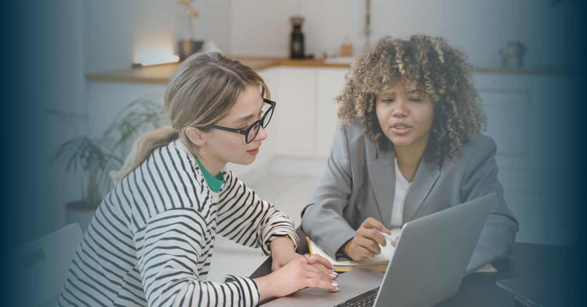 Two women sat a table looking at a laptop. One has long brown hair tied in a ponytail, thick black glasses and a stripey top, the other has curly brown hair and is wearing a light grey suit jacket.